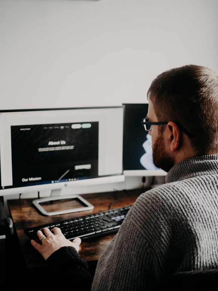 A man is working on web design from a home office setup with two monitors.
